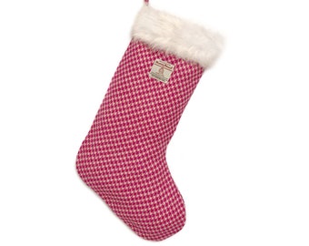 Könnte beinhalten: Ein Weihnachtsstrumpf mit einem rosa-weißen Hahnentrittmuster. Der Strumpf hat eine weiße Kunstfellmanschette und eine Aufhängeschlaufe. Ein kleines Etikett ist auf der Vorderseite angenäht.