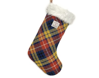 May include: A Christmas stocking featuring a vibrant red, yellow, and blue plaid pattern. The stocking has a white faux fur cuff at the top and a matching plaid hanging loop. A small white rectangular label with text is sewn onto the front.
