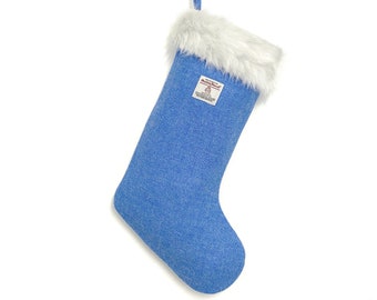 May include: A blue Christmas stocking made of textured fabric with a white faux fur cuff at the top. A blue fabric loop is attached for hanging. A small rectangular label with the text Harris Tweed is sewn onto the front of the stocking.