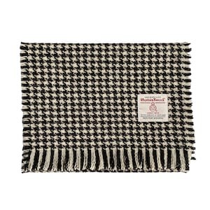 May include: A rectangular Harris Tweed scarf featuring a black and white houndstooth pattern. The fabric has a visible woven texture with frayed edges on all sides. A white rectangular label is sewn on the front with red and black text.