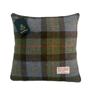 May include: Square cushion with a plaid pattern in shades of green, brown, and blue. The cushion has a tag that says "Harris Tweed". The cushion is a square shape.