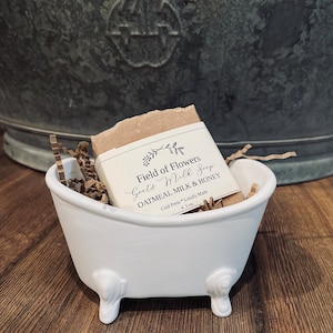 May include: A white ceramic bathtub-shaped soap dish with a bar of goat's milk soap inside. The soap is wrapped in brown paper and has a label that reads "Field of Flowers Goat's Milk Soap Oatmeal Milk & Honey Cold Press Locally Made 4.5 oz."