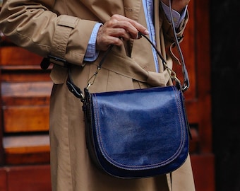 May include: A woman wearing a beige trench coat and blue jeans carries a dark blue leather satchel bag with a brown strap.
