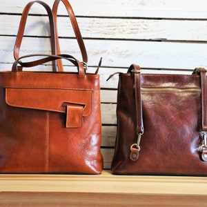 Pode incluir: Duas bolsas de couro em tons de castanho. A bolsa da esquerda é castanho claro com fecho de aba e alças superiores. A bolsa da direita é castanho escuro com fecho de correr e alça de ombro. Ambas as bolsas estão sobre uma superfície de madeira.