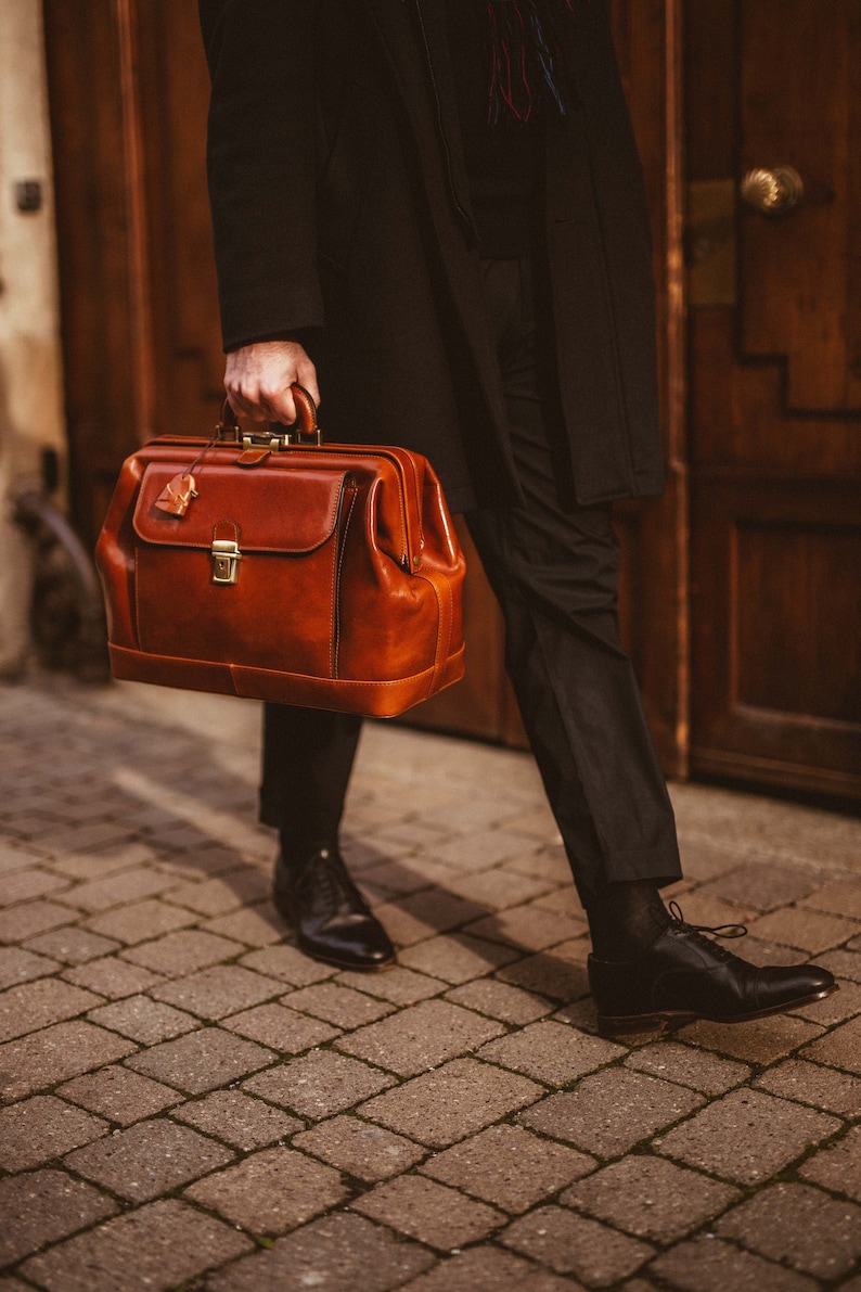 May include: A brown leather doctor's bag with a top handle and a clasp closure. The bag has a rectangular shape and a small tag attached. The person is wearing black pants and shoes.