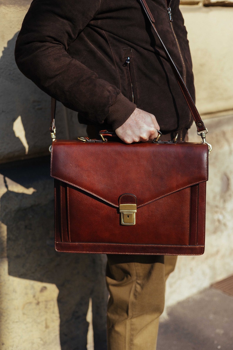 Pu&ograve; includere: Una ventiquattrore rettangolare in pelle marrone scuro con finitura liscia, manico superiore e tracolla rimovibile. La patta frontale &egrave; fissata con una chiusura a scatto in metallo color ottone e presenta dettagli con cuciture verticali.