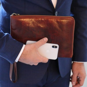 May include: A brown leather clutch with a zip closure and a gold zip pull. The clutch is being held in the hand of a person wearing a blue suit.