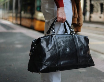 May include: A black leather duffel bag with silver hardware is held by a person wearing a red sweater and white pants. The bag has two top handles and a shoulder strap. The person is wearing white and beige sneakers. A brown jacket is draped over the person's arm.