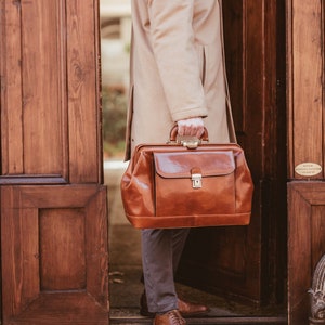 May include: A brown leather doctor's bag with a top handle and a metal clasp. The bag is held by a person wearing a tan overcoat and gray pants. The person is standing in front of a wooden door.