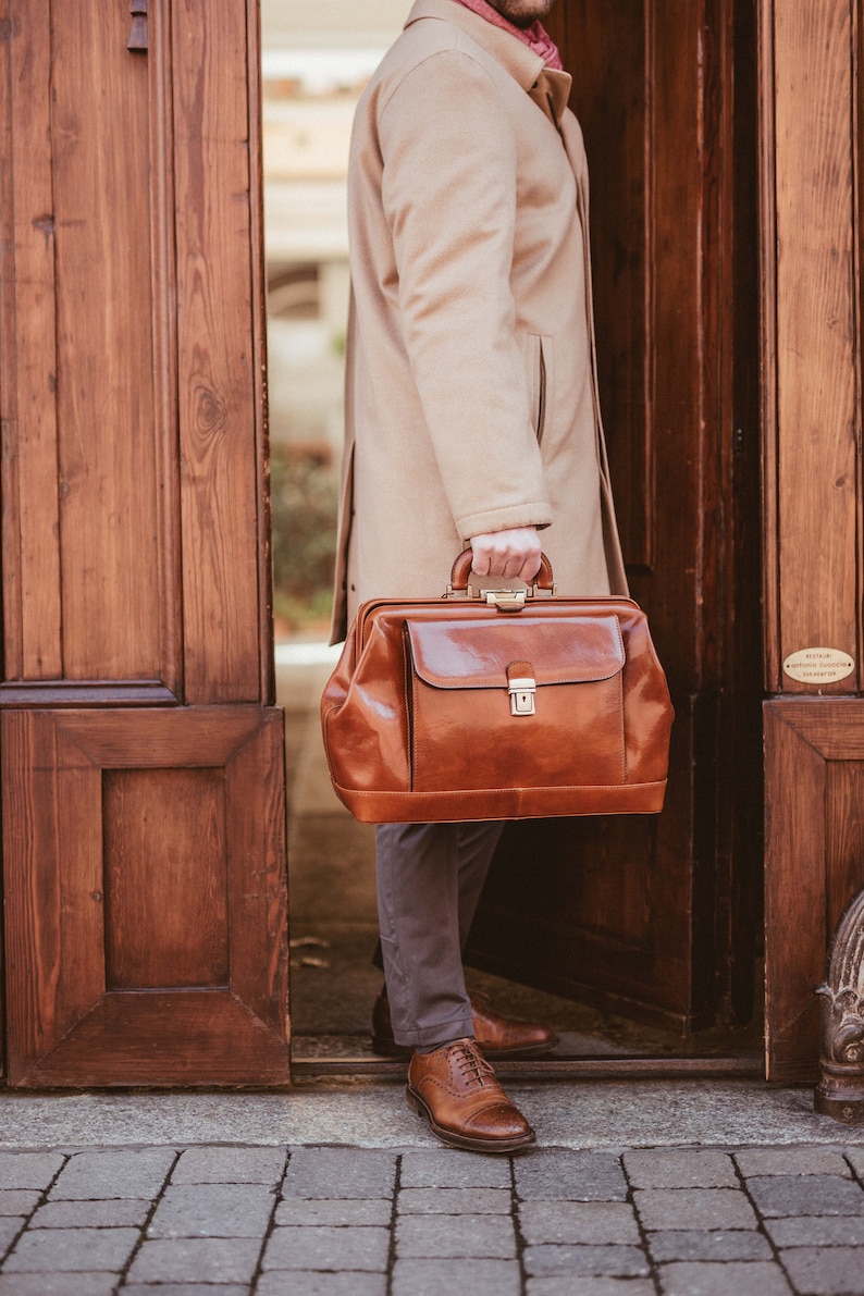 May include: A brown leather doctor's bag with a top handle and a metal clasp. The bag is held by a person wearing a tan overcoat and gray pants. The person is standing in front of a wooden door.