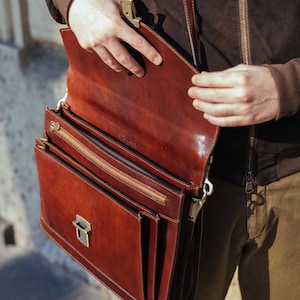 Pu&ograve; includere: Una cartella in pelle marrone con silhouette rettangolare, patta con chiusura metallica e tracolla regolabile. L'interno presenta uno scomparto con cerniera e cuciture visibili lungo i bordi della pelle.