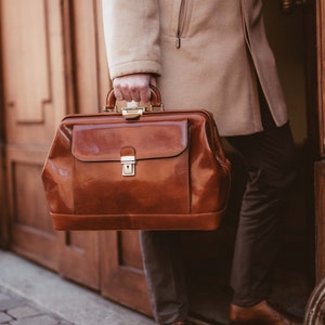 May include: A brown leather doctor's bag with a shiny finish, held by a person wearing a tan coat. The bag has a rectangular shape with a top handle and a front flap secured by a gold-colored clasp. The person is wearing brown shoes.