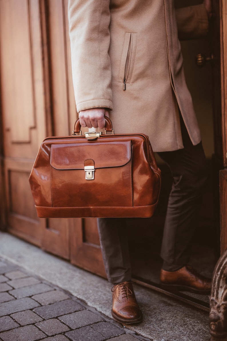 May include: A brown leather doctor's bag with a shiny finish, held by a person wearing a tan coat. The bag has a rectangular shape with a top handle and a front flap secured by a gold-colored clasp. The person is wearing brown shoes.