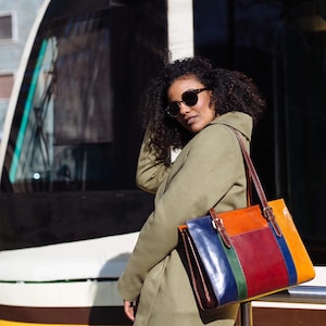 May include: A woman wearing a green coat and sunglasses stands next to a yellow and white train. She is carrying a multi-colored leather tote bag with brown straps.