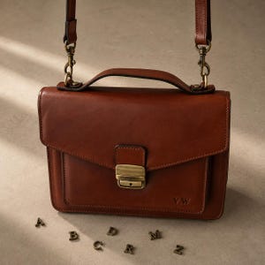 Op de afbeelding: Een bruine leren tas met een handvat aan de bovenkant, een afneembare schouderriem met messingkleurige draaibare clips en een voorflap die wordt vastgezet met een messingkleurige metalen sluiting. De initialen V.W. staan in de voorzak gedrukt.