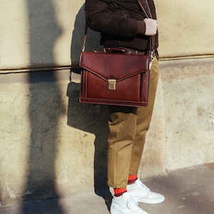 Pu&ograve; includere: Una ventiquattrore in pelle marrone scuro caratterizzata da una forma rettangolare strutturata, un manico superiore e una chiusura con fibbia in metallo dorato. La borsa include una tracolla regolabile e cuciture visibili lungo i bordi.