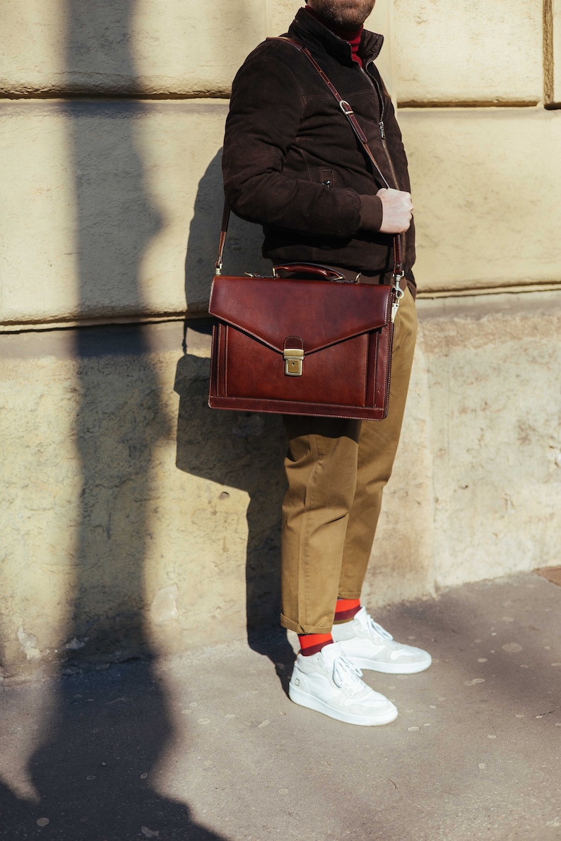 Pu&ograve; includere: Una ventiquattrore in pelle marrone scuro caratterizzata da una forma rettangolare strutturata, un manico superiore e una chiusura con fibbia in metallo dorato. La borsa include una tracolla regolabile e cuciture visibili lungo i bordi.