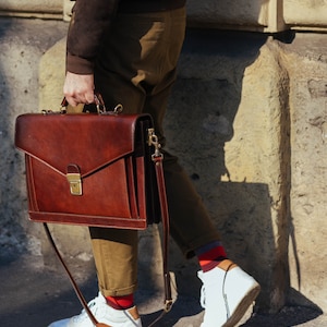 Pu&ograve; includere: Una ventiquattrore in pelle marrone scuro dalla forma rettangolare strutturata, con manico superiore e chiusura in metallo color ottone. La borsa presenta una tracolla rimovibile con finiture in ottone e cuciture visibili sui bordi.