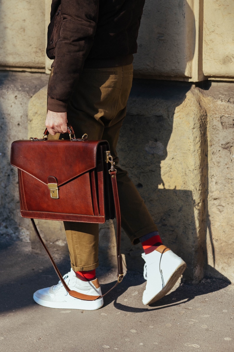 Pu&ograve; includere: Una ventiquattrore in pelle marrone scuro dalla forma rettangolare strutturata, con manico superiore e chiusura in metallo color ottone. La borsa presenta una tracolla rimovibile con finiture in ottone e cuciture visibili sui bordi.