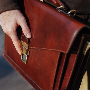 Pu&ograve; includere: Una cartella strutturata in pelle marrone dotata di manico superiore, patta frontale e chiusura a pressione in metallo color ottone. La borsa presenta cuciture visibili lungo i bordi e una finitura liscia e lucida con dettagli in marrone scuro.