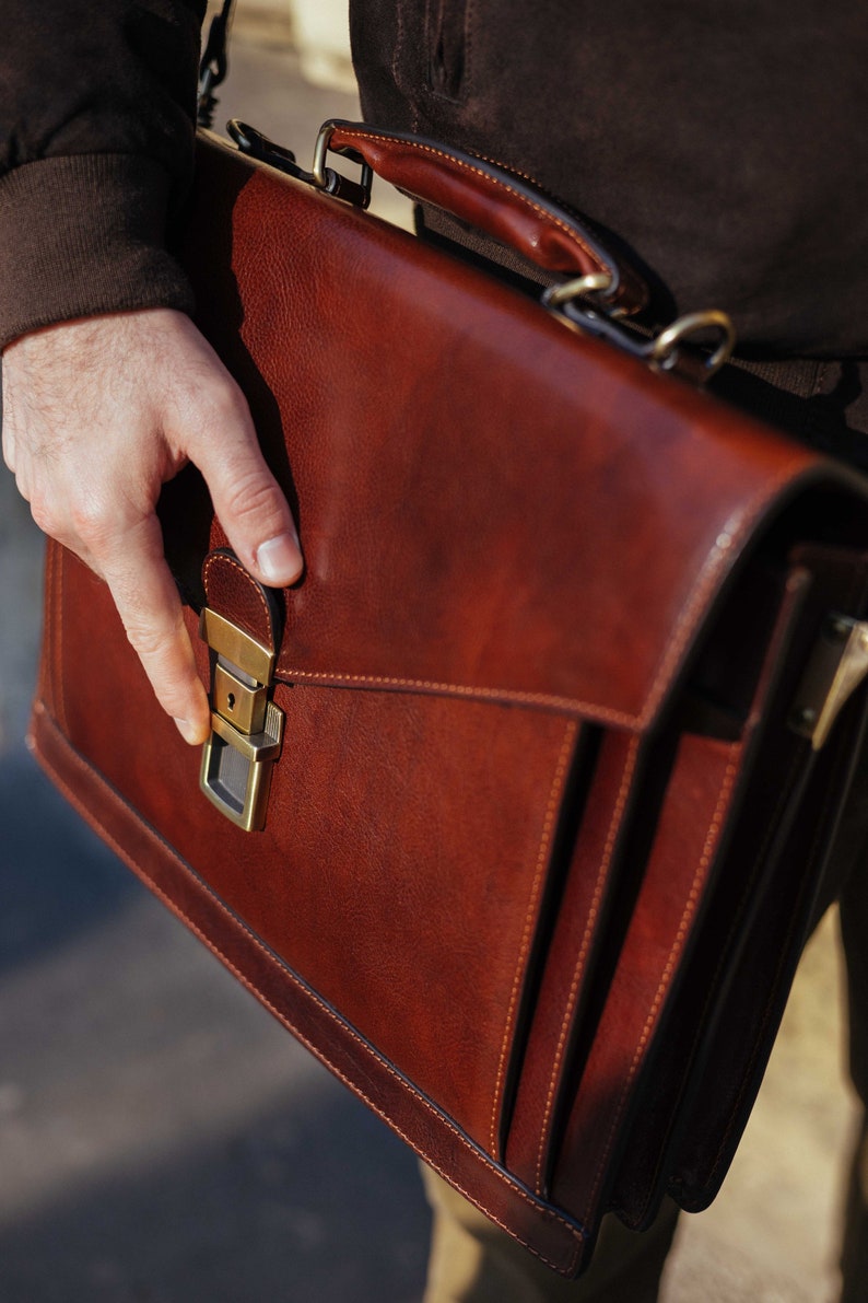 Pu&ograve; includere: Una cartella strutturata in pelle marrone dotata di manico superiore, patta frontale e chiusura a pressione in metallo color ottone. La borsa presenta cuciture visibili lungo i bordi e una finitura liscia e lucida con dettagli in marrone scuro.