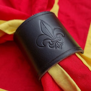 May include: A dark brown leather napkin ring featuring an embossed fleur-de-lis symbol. The ring is holding a bright red fabric napkin with a wide yellow border, creating a high-contrast display of colour and texture in this functional table accessory.