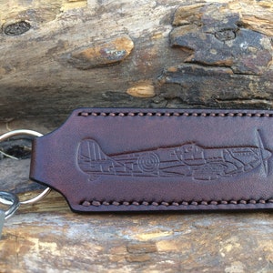 May include: Brown leather keyring with a stitched border and a debossed image of a vintage aircraft.