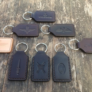 May include: Eight leather keychains with different designs, including a winged emblem, a dagger, crossed rifles, and a crest. The keychains are brown and black and have silver key rings.