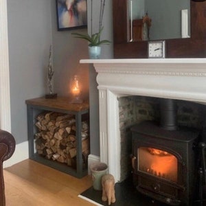 May include: A wood and metal log rack with stacked firewood in front of a white fireplace with a black wood-burning stove. The stove has a glass door and a glowing fire inside.