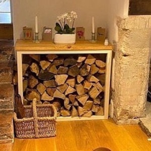 May include: A white wooden log rack with a square opening, filled with stacked firewood. The rack is sitting on a wooden floor in front of a fireplace.