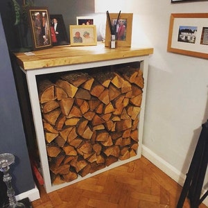 May include: A white wood log rack with a wooden top shelf. The rack is filled with stacked firewood and has a white frame. The top shelf has a few framed photos and a diffuser.