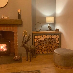 May include: A gray wood log rack with a metal frame, filled with stacked logs. A gray velvet ottoman sits in front of the rack. A deer statue stands to the left of the rack. A lamp with a white shade and a rabbit figurine sits on top of the rack. The word "Home" is written on a wooden beam above a fireplace.