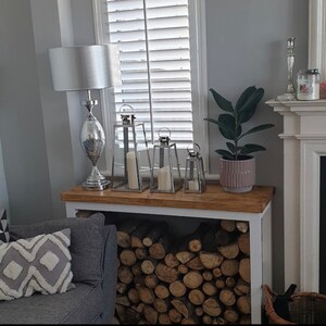 May include: A white wood log holder with a white top shelf. The shelf has a silver lamp with a white shade, three silver lanterns, and a potted plant with green leaves. The log holder is filled with stacked logs.