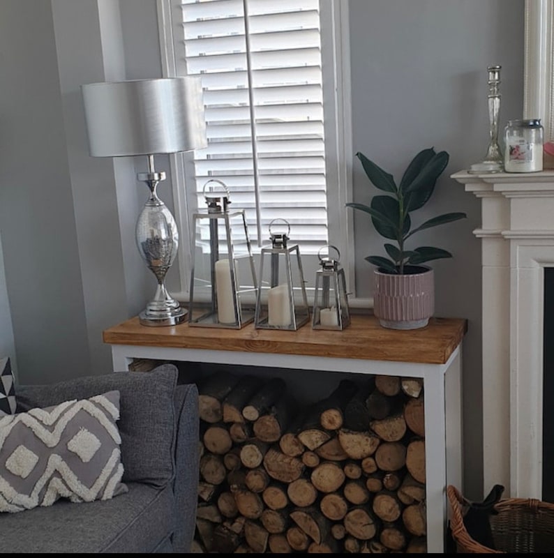 May include: A white wood log holder with a white top shelf. The shelf has a silver lamp with a white shade, three silver lanterns, and a potted plant with green leaves. The log holder is filled with stacked logs.