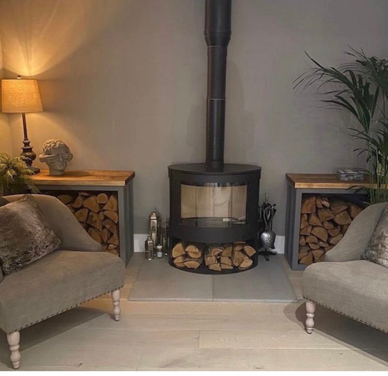 May include: A black wood-burning stove with a glass door and a metal chimney. The stove is surrounded by two wooden storage shelves with stacked firewood. The shelves are painted gray and have a light wood top.