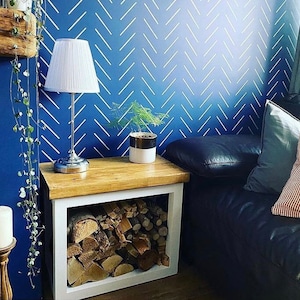 May include: A white wood log holder with a shelf on top. The log holder is filled with firewood. A white lamp with a white lampshade is on the shelf. A small potted plant is on the shelf next to the lamp. The log holder is in front of a blue wall with a white chevron pattern.