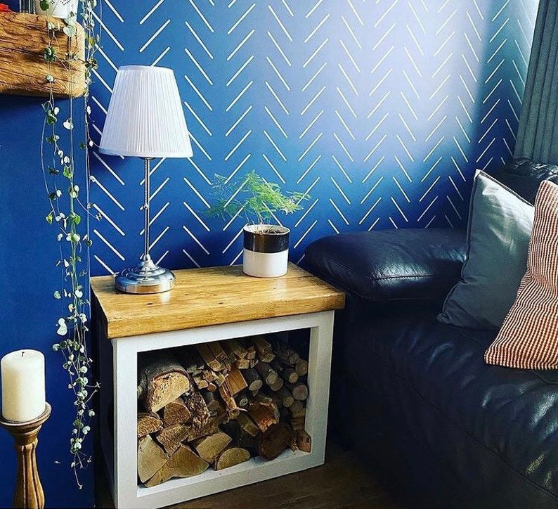 May include: A white wood log holder with a shelf on top. The log holder is filled with firewood. A white lamp with a white lampshade is on the shelf. A small potted plant is on the shelf next to the lamp. The log holder is in front of a blue wall with a white chevron pattern.