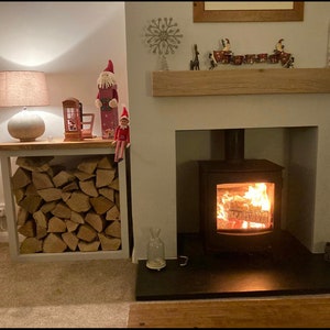 May include: A white wood log rack with stacked firewood sits next to a fireplace with a black wood-burning stove. The stove has a glass door and a roaring fire inside. A white mantel above the fireplace is decorated with a wooden snowflake, reindeer, and a row of small red and white ornaments.