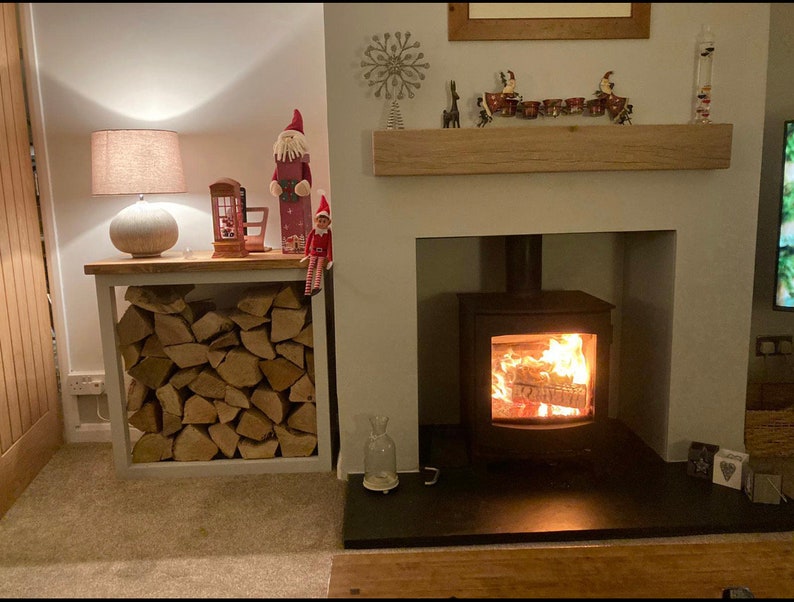 May include: A white wood log rack with stacked firewood sits next to a fireplace with a black wood-burning stove. The stove has a glass door and a roaring fire inside. A white mantel above the fireplace is decorated with a wooden snowflake, reindeer, and a row of small red and white ornaments.
