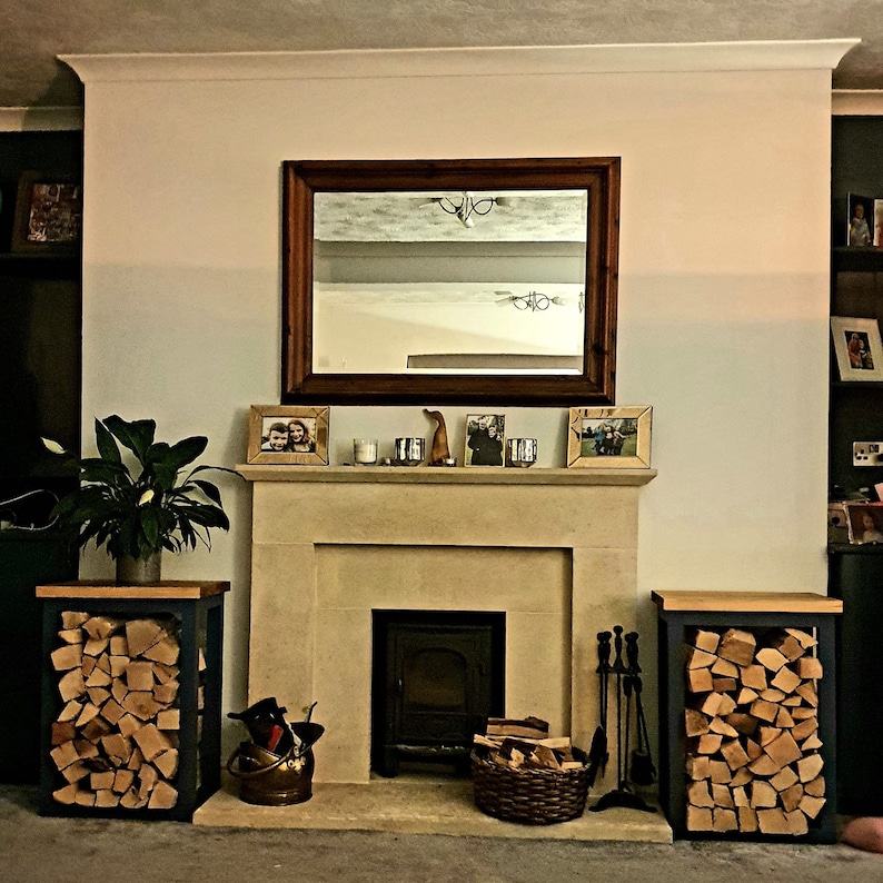 May include: A fireplace with a wood-burning stove and a mantelpiece decorated with framed photos and a small wooden figurine. Two black log racks with stacked firewood are on either side of the fireplace.