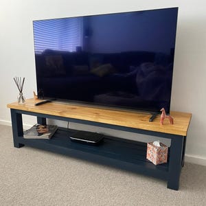 May include: A wooden TV stand with a natural wood top and navy blue base. A large flat-screen TV sits on top. Decorative items include a small giraffe figurine, a vase with reeds, and a patterned tissue box.