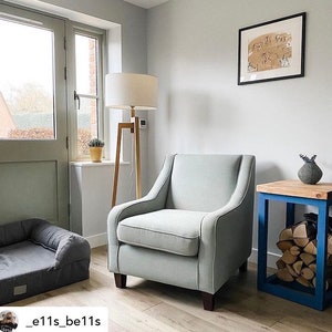 May include: A light gray armchair with a wooden frame sits in a living room with a wooden floor. A small wooden table with a blue frame holds a vase with dried flowers. A dog bed is in the foreground.