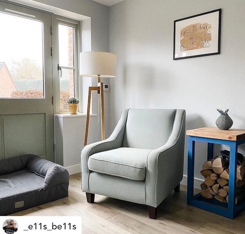 May include: A light gray armchair with a wooden frame sits in a living room with a wooden floor. A small wooden table with a blue frame holds a vase with dried flowers. A dog bed is in the foreground.
