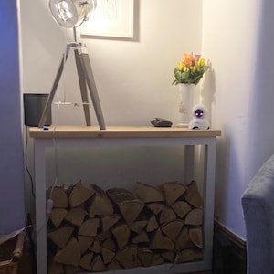 May include: A white wooden log rack with a stack of firewood inside. A silver tripod lamp with a spotlight shade sits on top of the rack. A small white robot and a vase of flowers are on the top shelf of the rack.