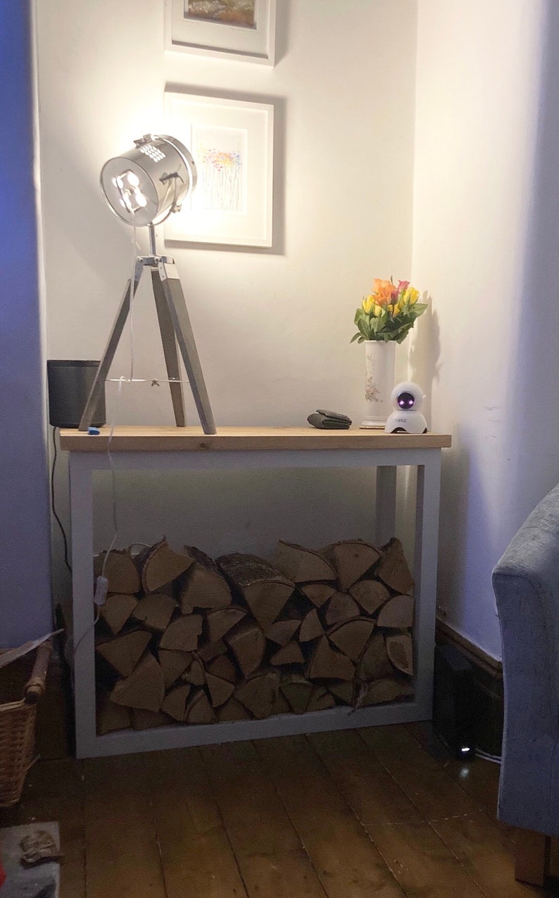 May include: A white wooden log rack with a stack of firewood inside. A silver tripod lamp with a spotlight shade sits on top of the rack. A small white robot and a vase of flowers are on the top shelf of the rack.