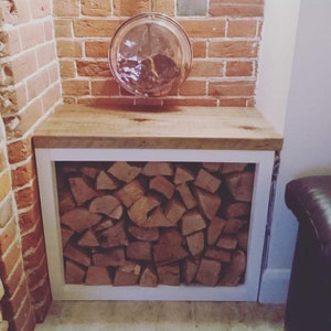 May include: A white wood log rack with a wooden top shelf. The rack is filled with stacked firewood and is set against a brick wall.