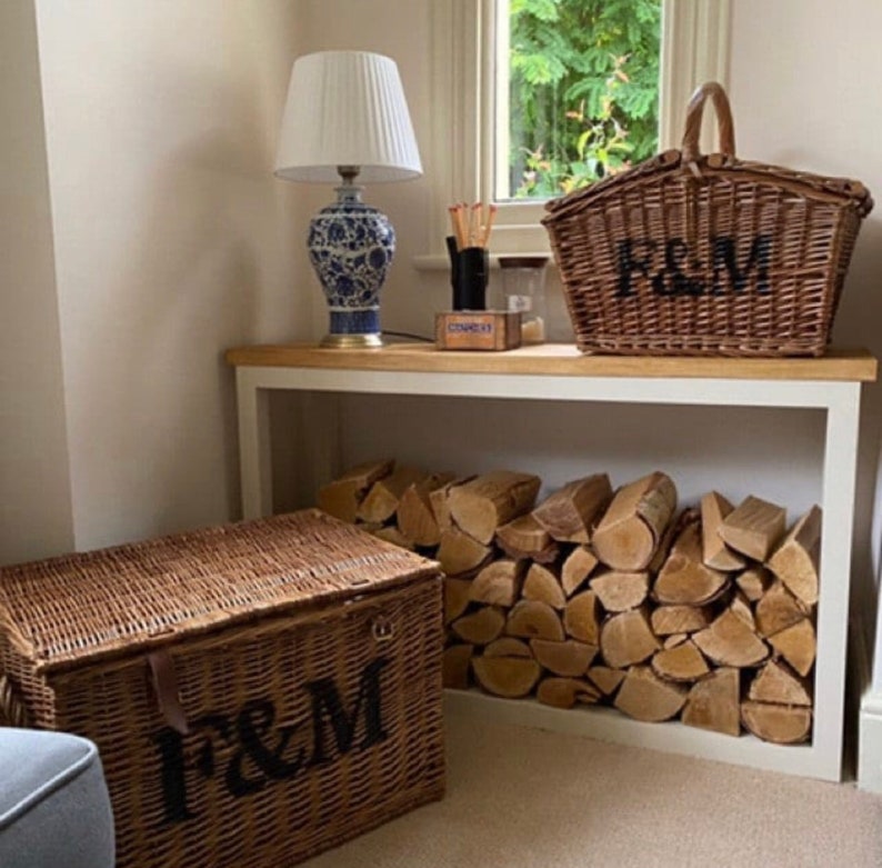 May include: Two wicker baskets with the letters "F&M" on them. The baskets are sitting on a white shelf with a stack of firewood behind them. A lamp with a white shade and a blue and white ceramic base is on the shelf above the baskets.