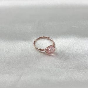 Puede incluir: Un delicado anillo de oro rosa con una piedra preciosa rosa iridiscente. El anillo est&aacute; hecho de alambre envuelto alrededor de la piedra.