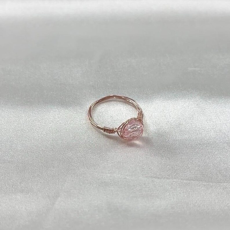Puede incluir: Un delicado anillo de oro rosa con una piedra preciosa rosa iridiscente. El anillo est&aacute; hecho de alambre envuelto alrededor de la piedra.