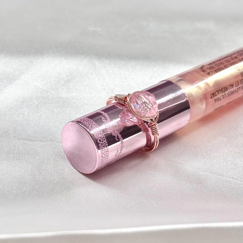 Puede incluir: Un anillo rosa y oro rosa con una piedra transparente iridiscente. El anillo est&aacute; envuelto en alambre y est&aacute; sobre una botella rosa.